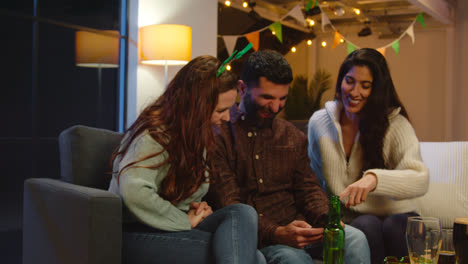 Group-Of-Friends-Dressing-Up-At-Home-Or-In-Bar-Celebrating-At-St-Patrick's-Day-Party-Looking-At-Photos-On-Phone